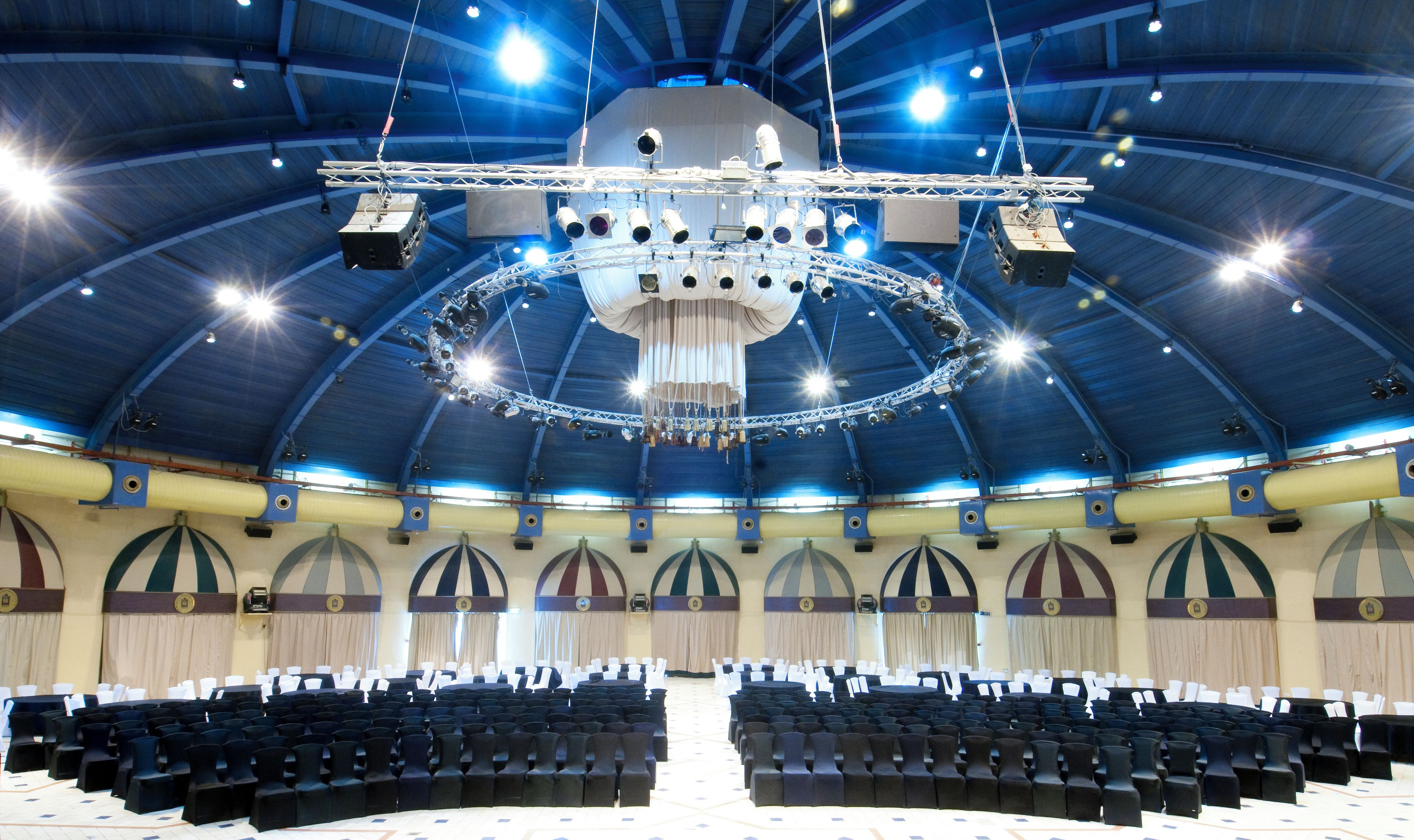 A large hall with rows of chairs, a stage with lighting equipment, and a high ceiling with a circular light fixture.