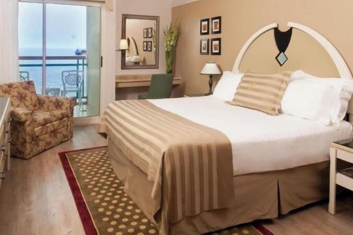 A hotel room with a bed, a chair, a desk, and a view of the sea.