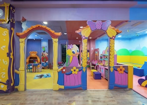 Zona de juegos infantiles con murales coloridos, un gran oso de peluche y una mesa de juegos.