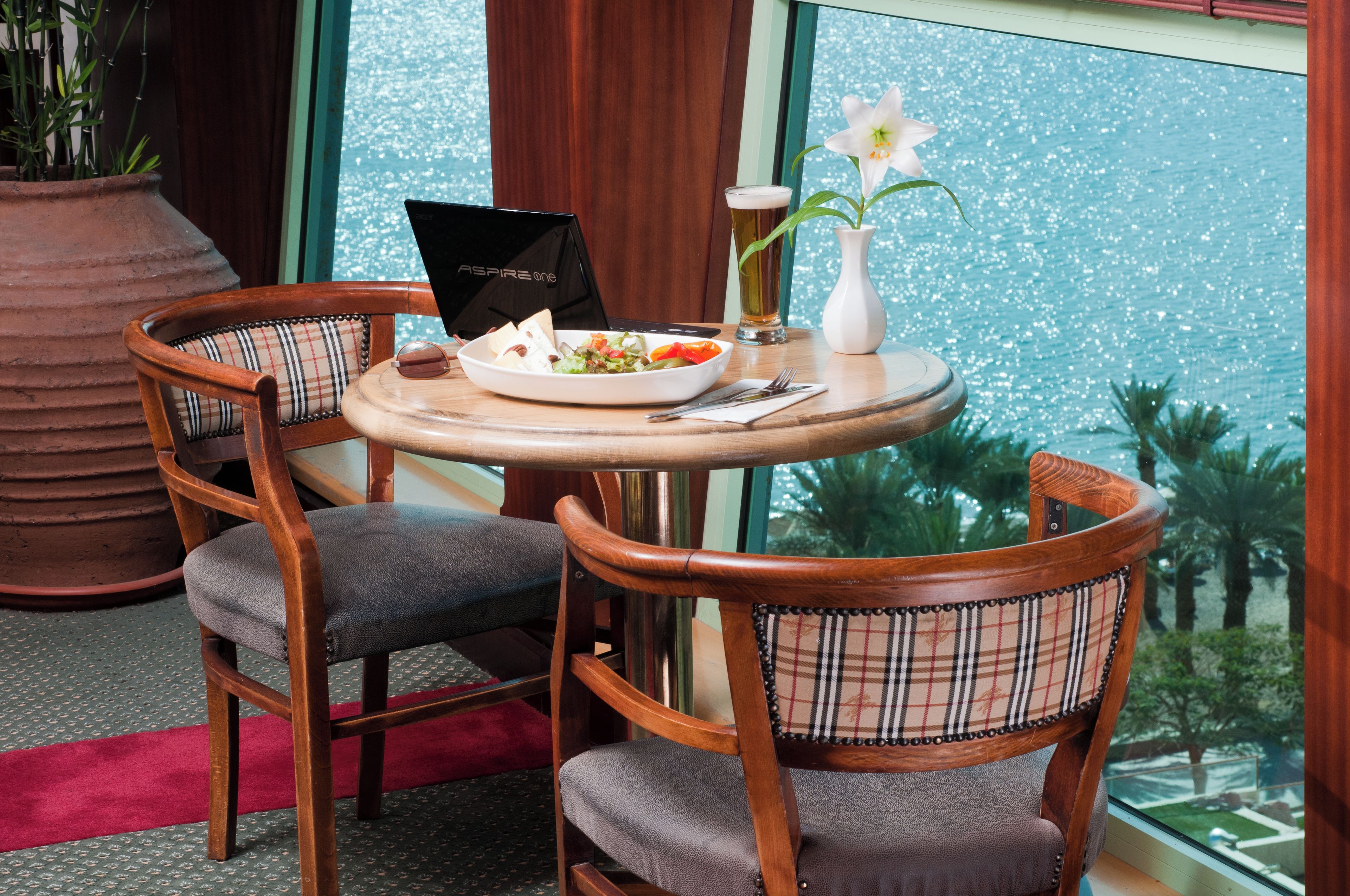 A dining area with two wooden chairs, a round table, a laptop, a bowl of food, a glass of beer, and a vase with a flower.