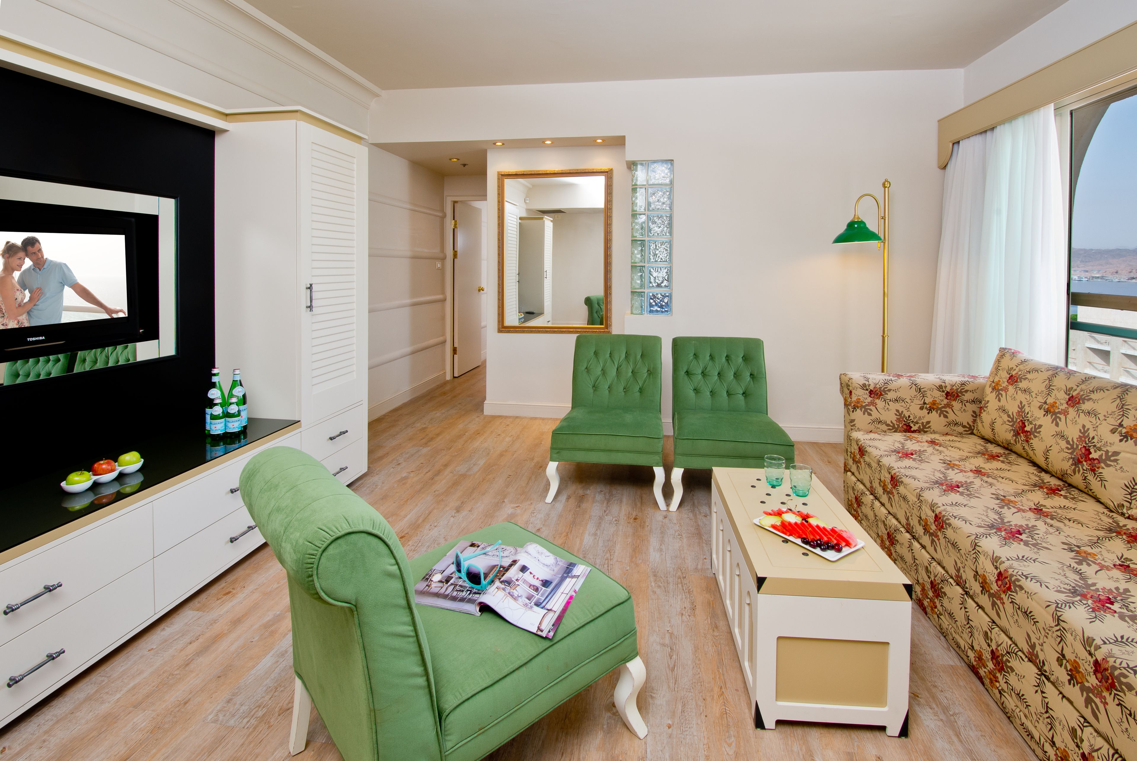 A modern living room with a flat-screen TV, green armchairs, a sofa, and a small table with drinks.