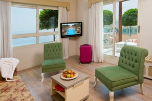 A hotel room with two green armchairs, a small table with fruit, a pink suitcase, and a TV displaying a pool view. - Herods Palace Hotels & Spa Eilat (Eilat)