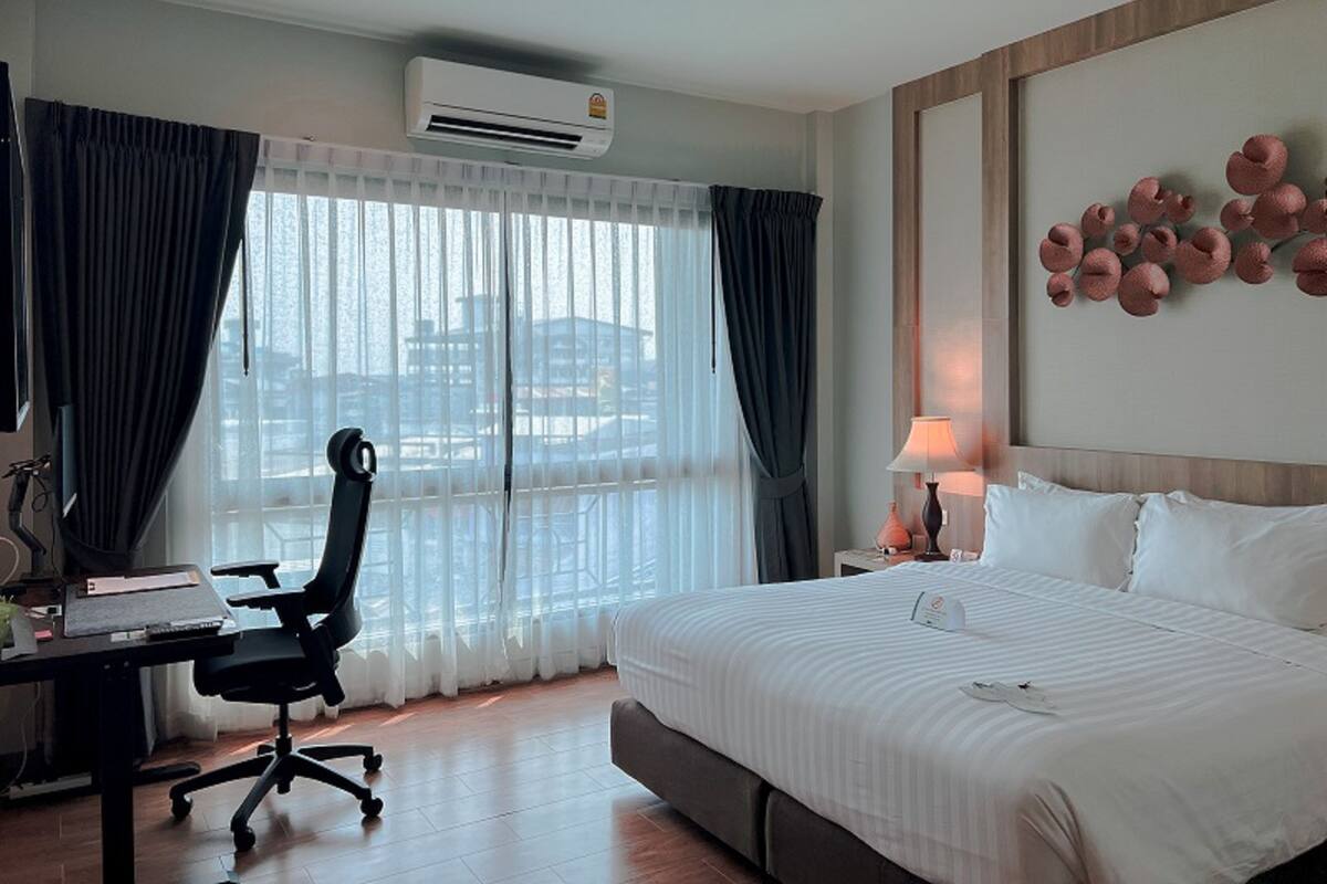 A hotel room with a large bed, a desk with a chair, an air conditioner, and a view of buildings through the window.
