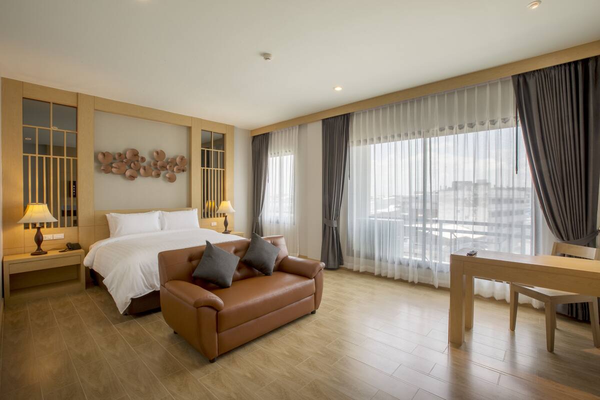 A hotel room with a bed, a brown sofa, a wooden table, and a large window with curtains.