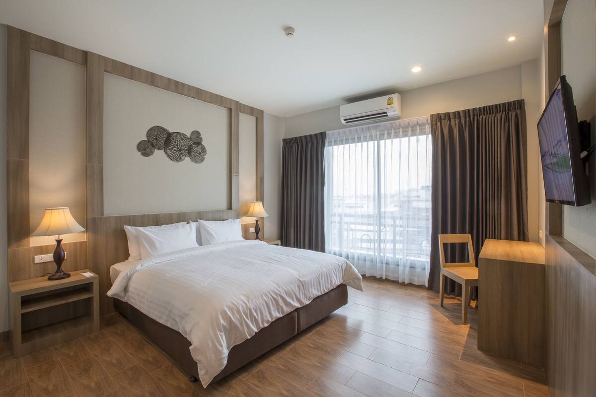 A modern hotel room with a large bed, two bedside tables with lamps, a flat-screen TV mounted on the wall, and a window with curtains.