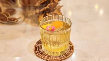 Un vaso de líquido color ámbar con una rodaja de naranja y una flor rosa como adorno sobre un posavasos tejido.