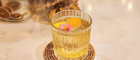 Un verre contenant un liquide de couleur ambrée, agrémenté d’une tranche d’orange et d’une fleur rose, posé sur un sous-verre en fibres tressées.