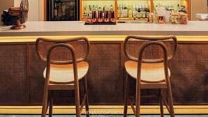 A bar area with a wooden ceiling, hanging light bulbs, and shelves stocked with bottles. There are two wicker chairs in front of the bar. - Civilize Hotel (Udon Thani)