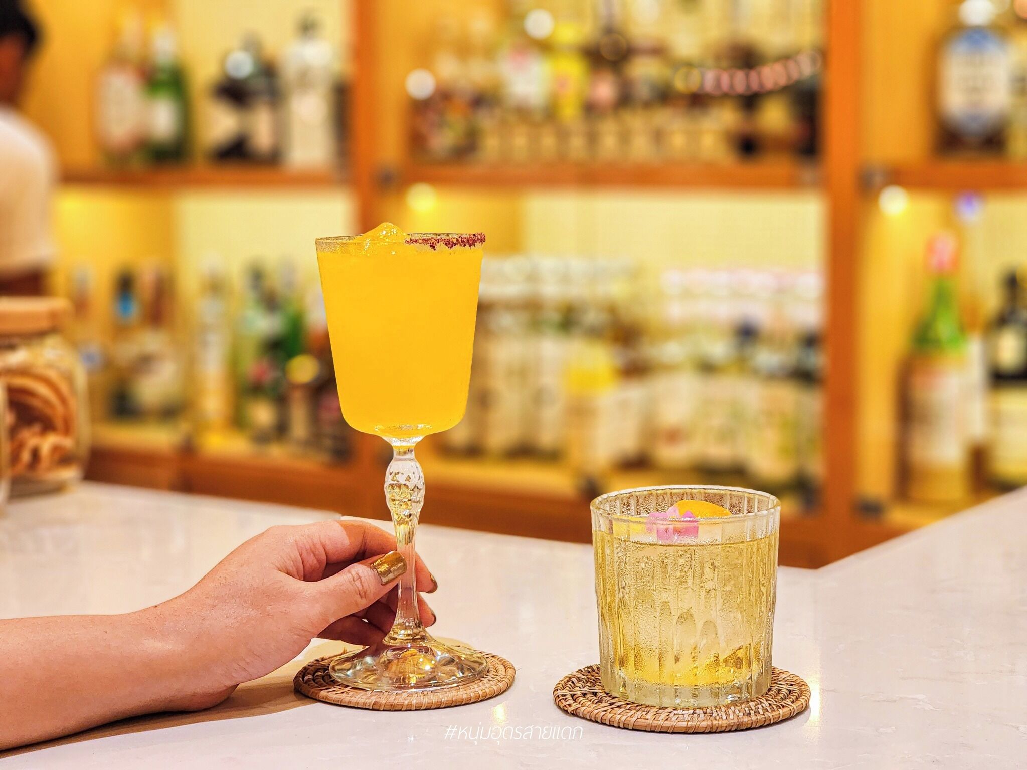 Un ambiente da bar con una mano che sorregge un bicchiere da cocktail e un bicchiere grande pieno di una bevanda dorata su un sottobicchieri.