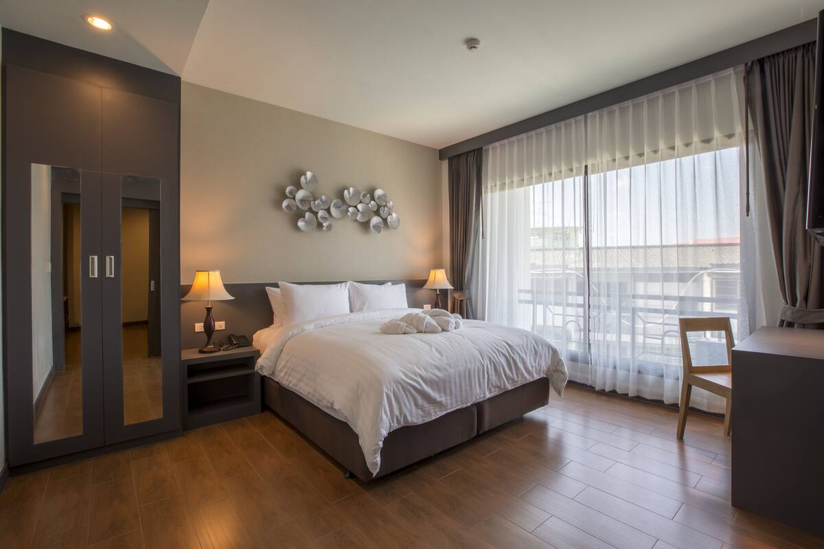 A modern hotel room with a large bed, bedside lamps, a desk, and a view of the city through large windows.