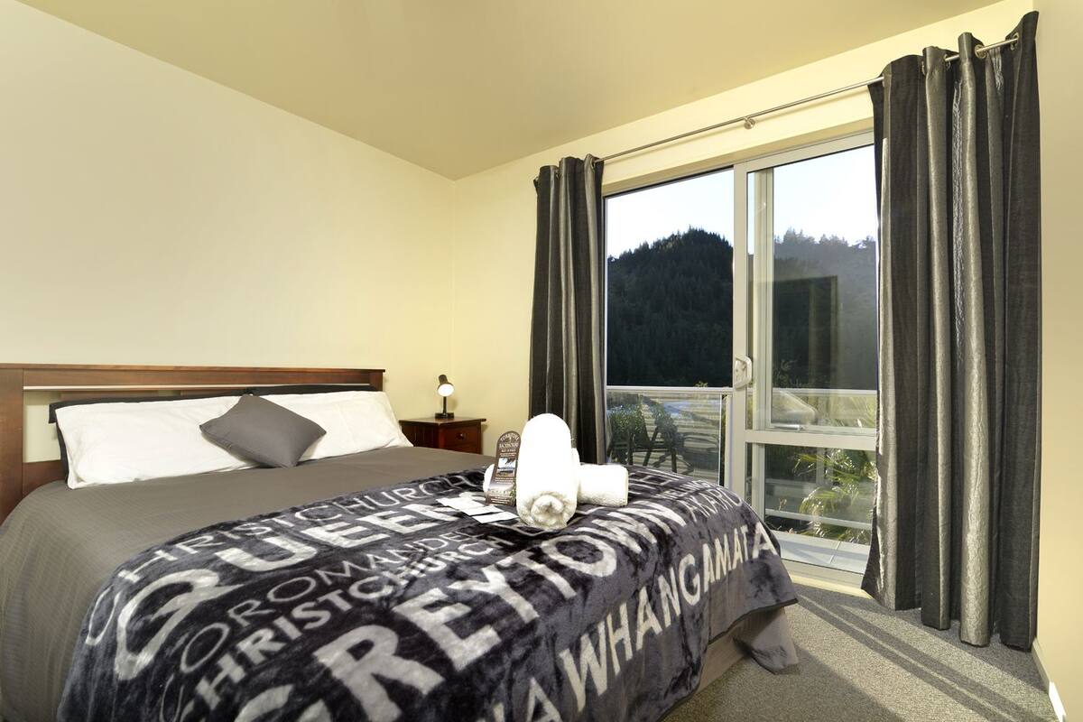 Habitación de hotel con cama, mesita de noche, ventana con cortinas y vista a árboles.