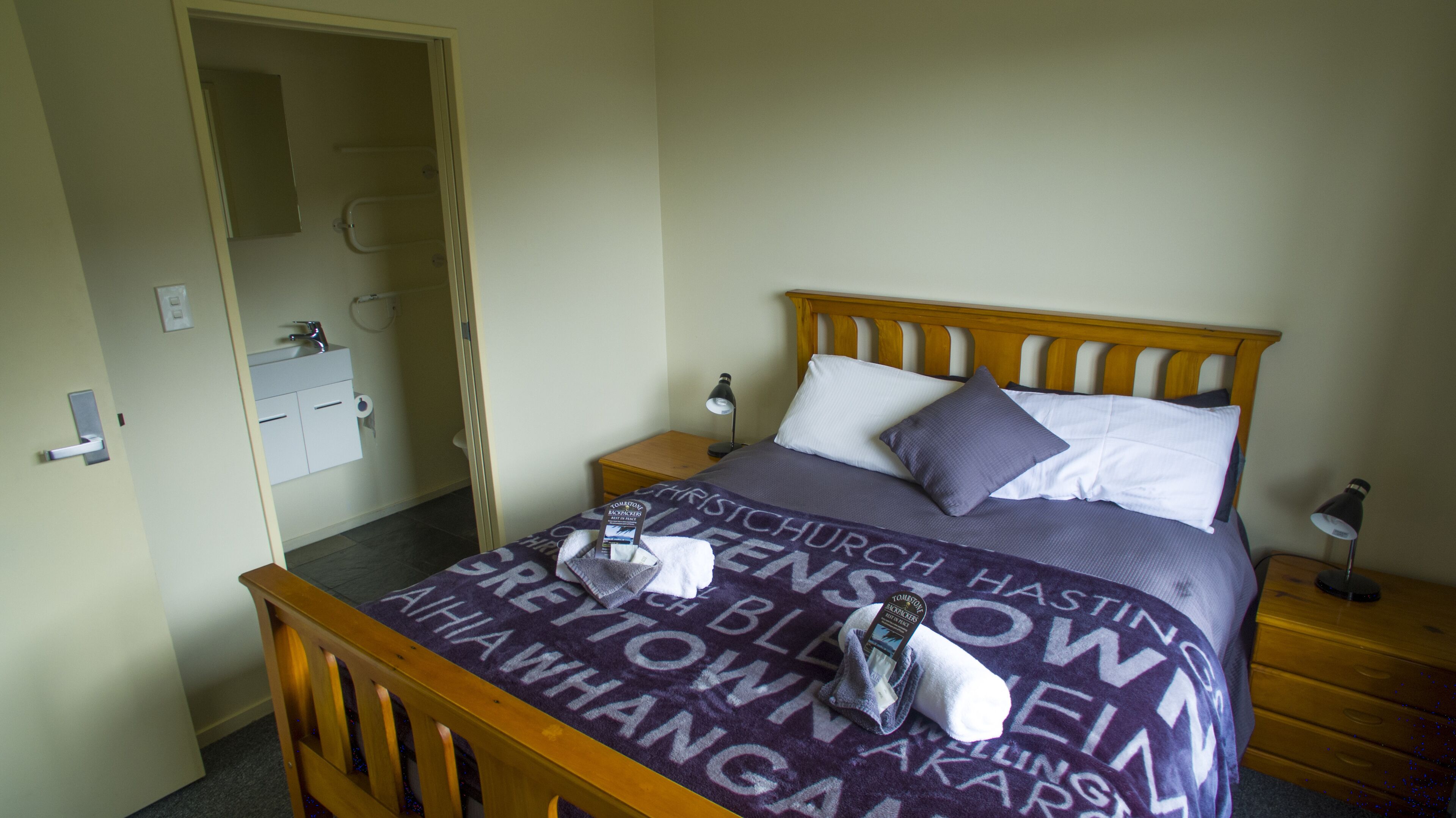 A bedroom with a wooden bed, a nightstand, a bedside lamp, a bathroom with a toilet, and a door.