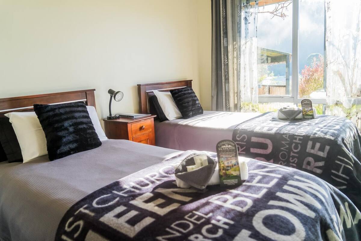 Habitación de hotel con dos camas, cada una con cabecera y almohadas, una mesita de noche con lámpara, y ventana con vista al exterior.