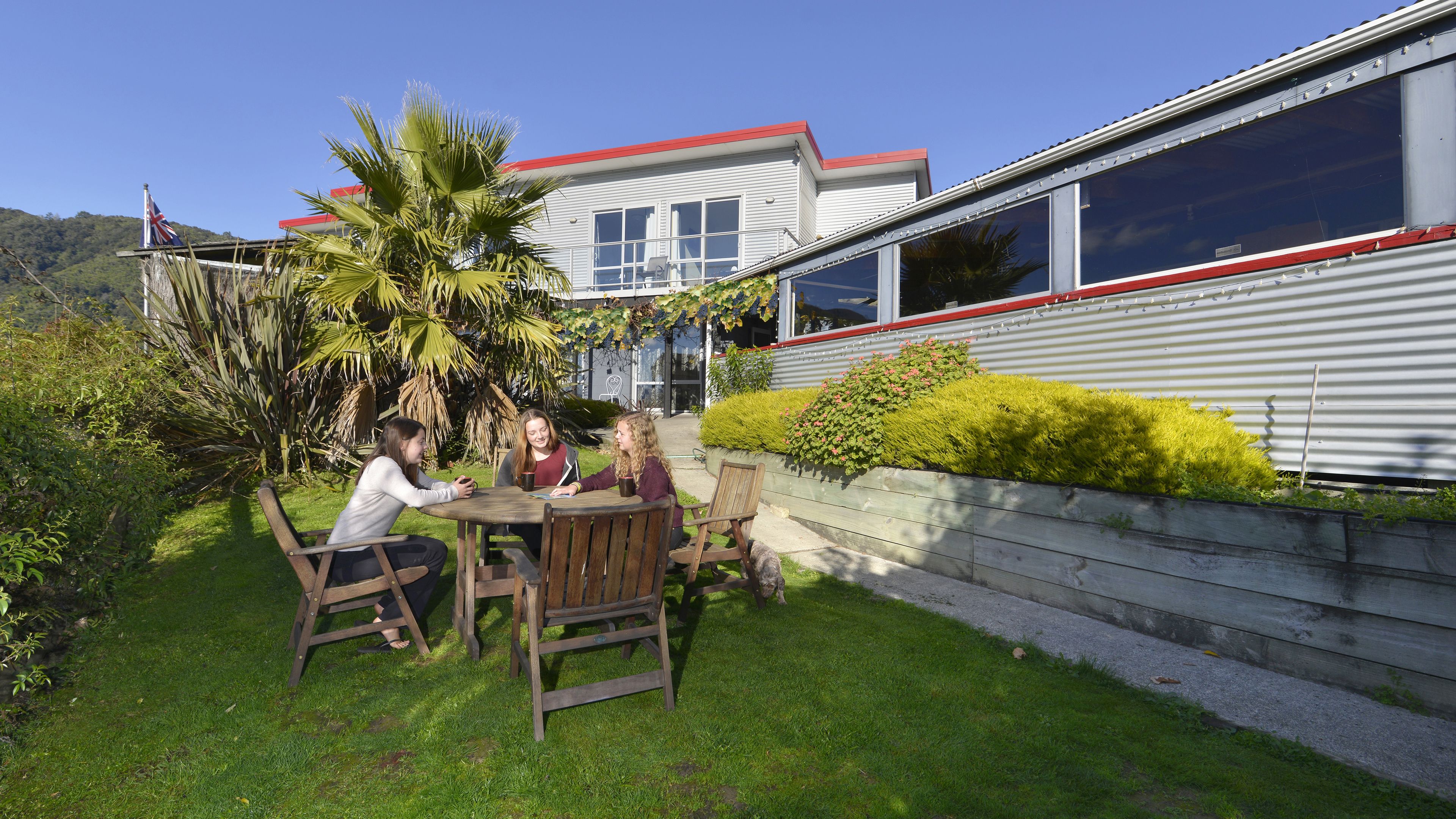 An outdoor seating area with a table and chairs, surrounded by greenery and a building with large windows.