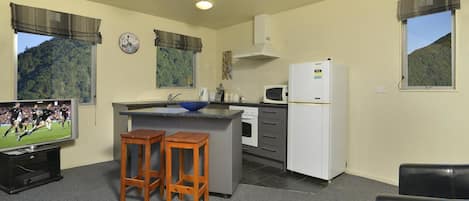 A compact kitchen with a sink, stove, microwave, and refrigerator. There are two wooden stools and a television displaying a sports event.