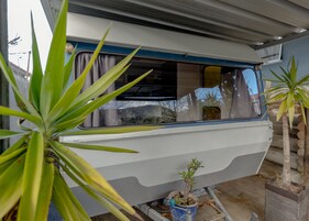 Una caravana blanca con una franja azul, una pequeña planta en maceta en el suelo y una ventana que refleja un edificio y árboles.