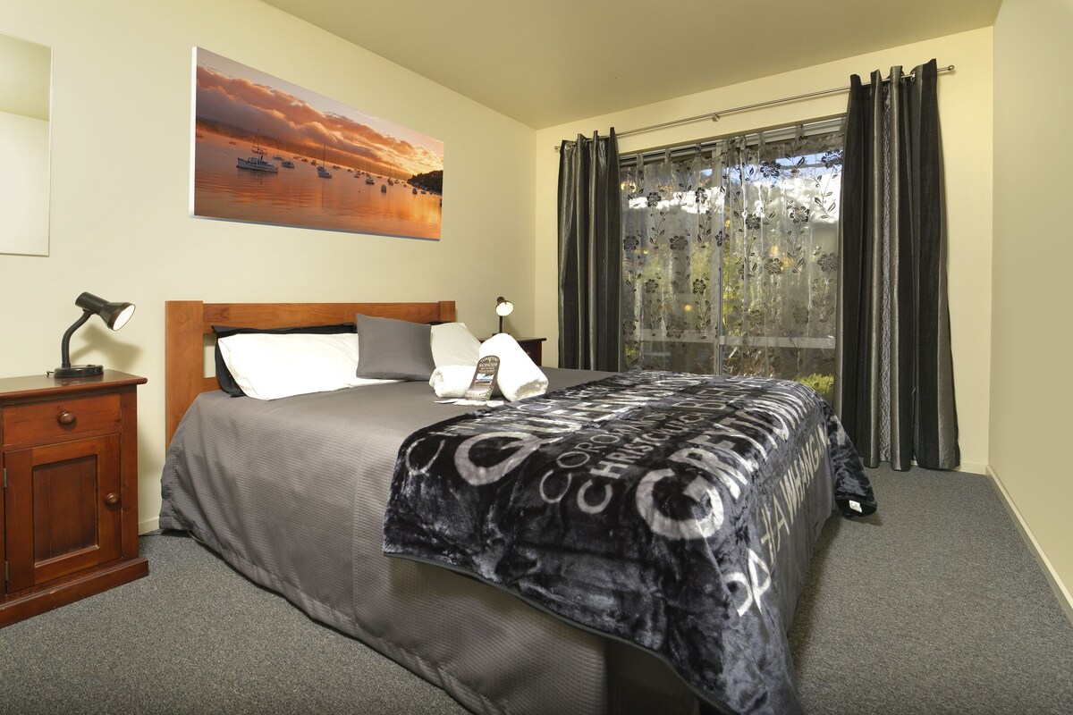 A neatly made bed with a patterned bedspread, a wooden nightstand with a lamp, a painting of a sunset over water on the wall, and a window with curtains.