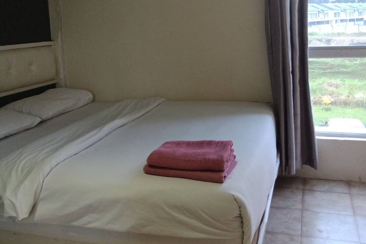 A single bed with white bedding and a folded red towel, a window with curtains, and a view of a building outside.
