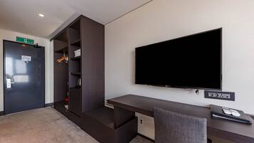 Une chambre d’hôtel moderne avec un bureau en bois foncé, un téléviseur à écran plat, un placard intégré et un petit coin salon.