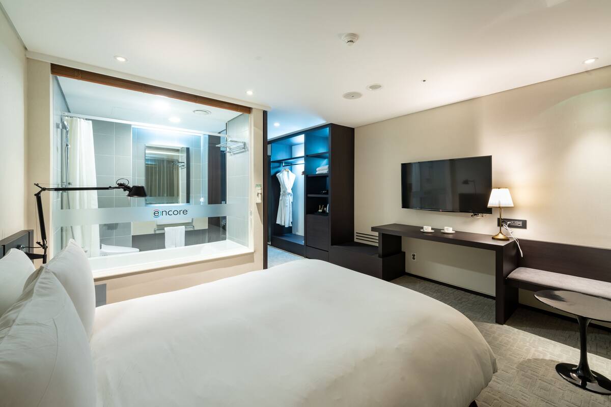 A modern hotel room with a large bed, a desk, a television, and a walk-in closet.