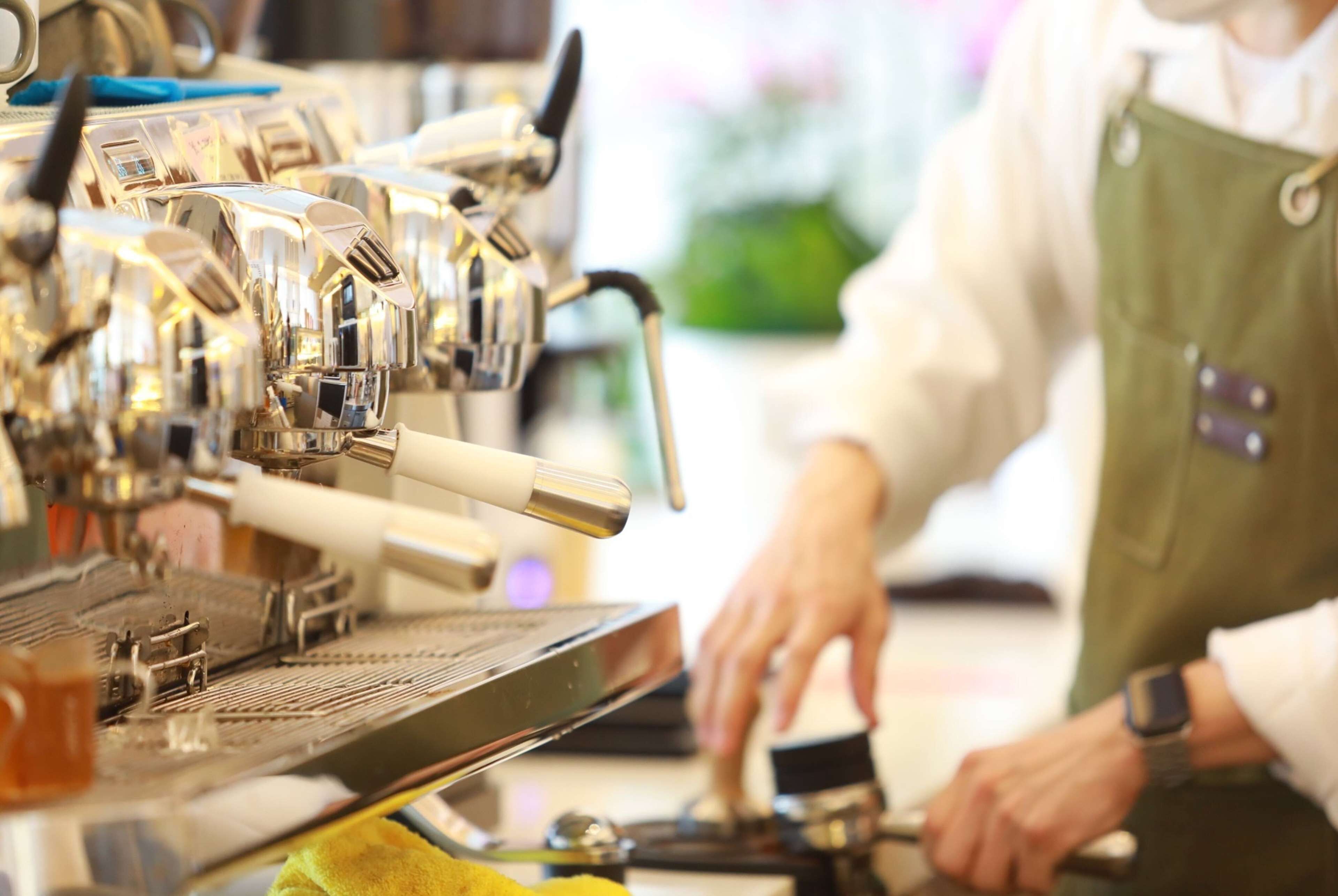 A close-up of a coffee machine with multiple dispensers, a person wearing a green apron, and a yellow cloth on the counter.