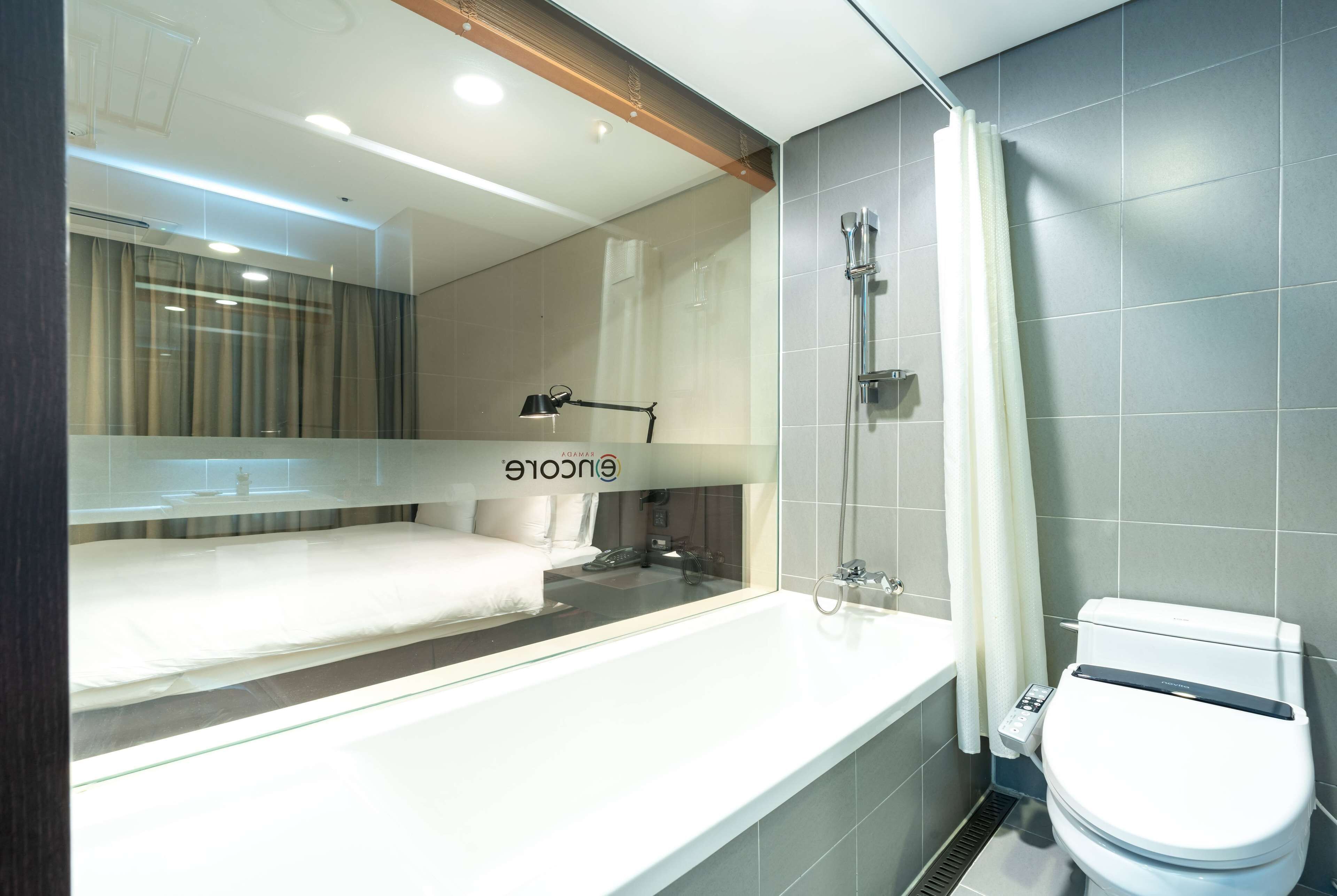 Un baño moderno con bañera, inodoro y un espejo grande que refleja una cama y cortinas.
