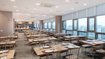 A spacious dining area with multiple tables and chairs, large windows offering a city view, and a modern kitchen in the background.