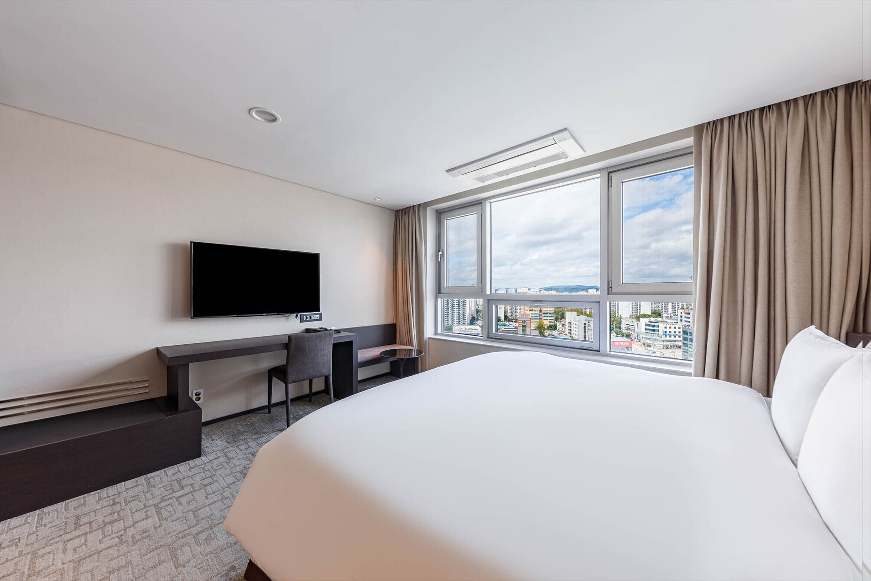 Habitación de hotel con una cama grande, un escritorio con silla, televisor de pantalla plana montado en la pared y un ventanal con vistas a la ciudad.