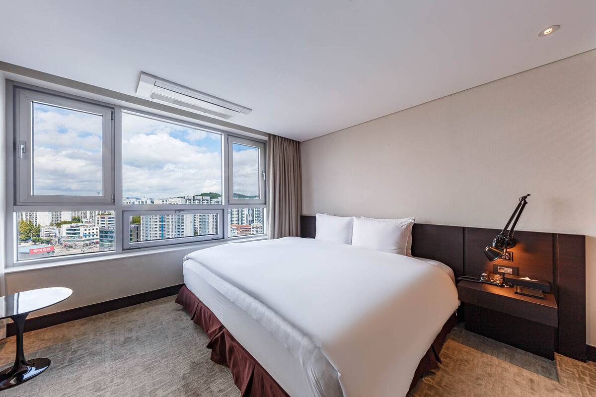A hotel room with a large bed, a bedside table with a lamp, a small table, and a view of the city through large windows.