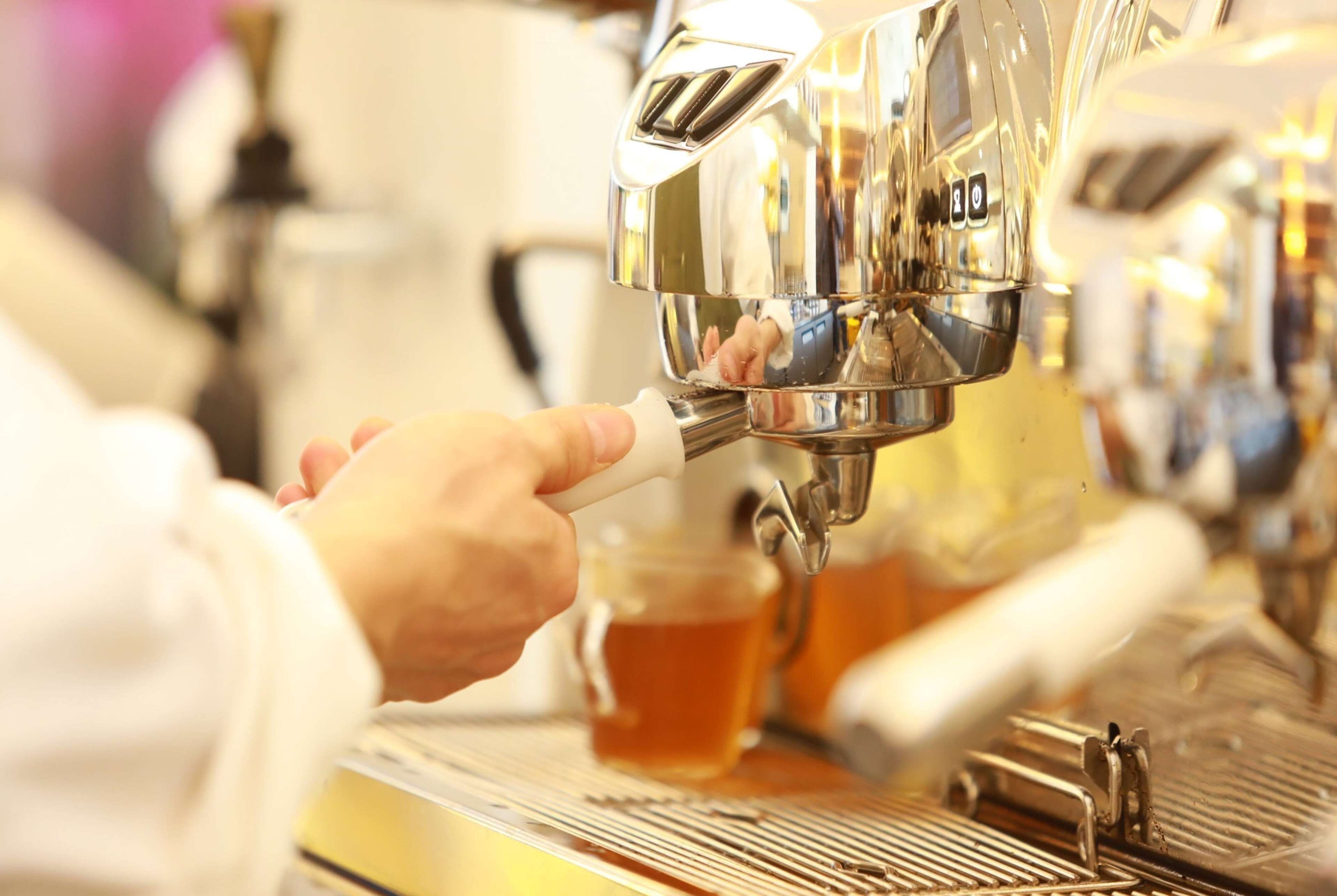 Gros plan d’une main manipulant une machine à café, avec une tasse de café en arrière-plan.