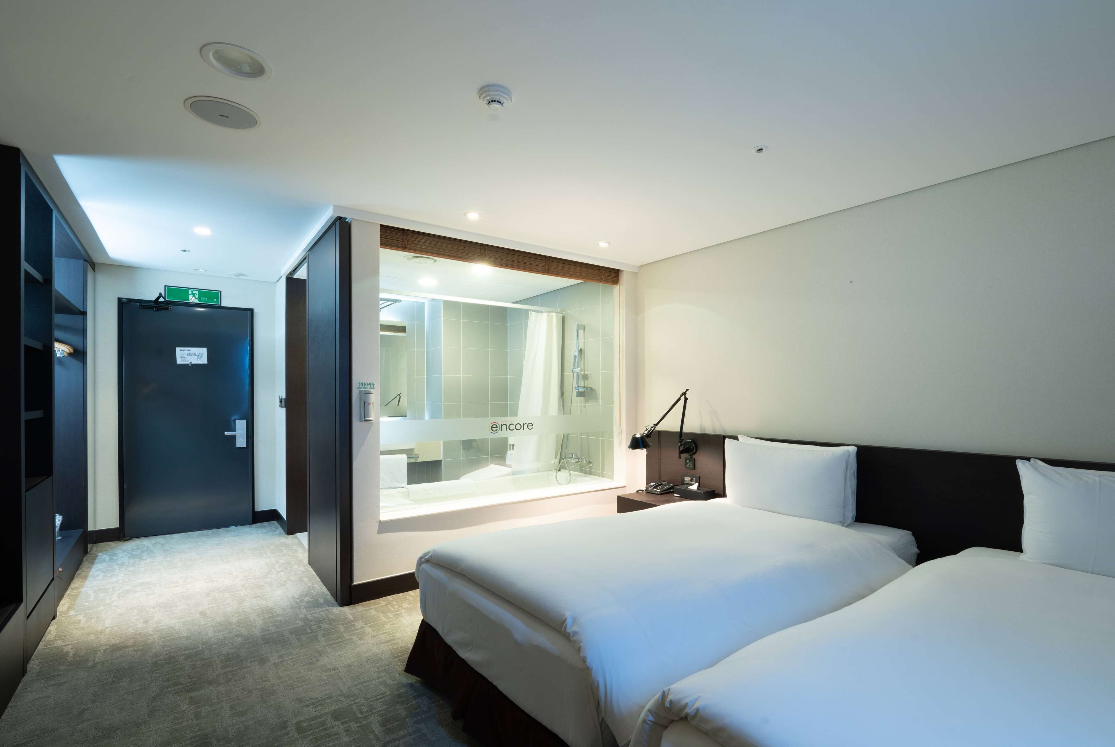 A hotel room with a large bed, a bedside table with a lamp, a bathroom with a glass shower enclosure, and a door with an emergency exit sign.