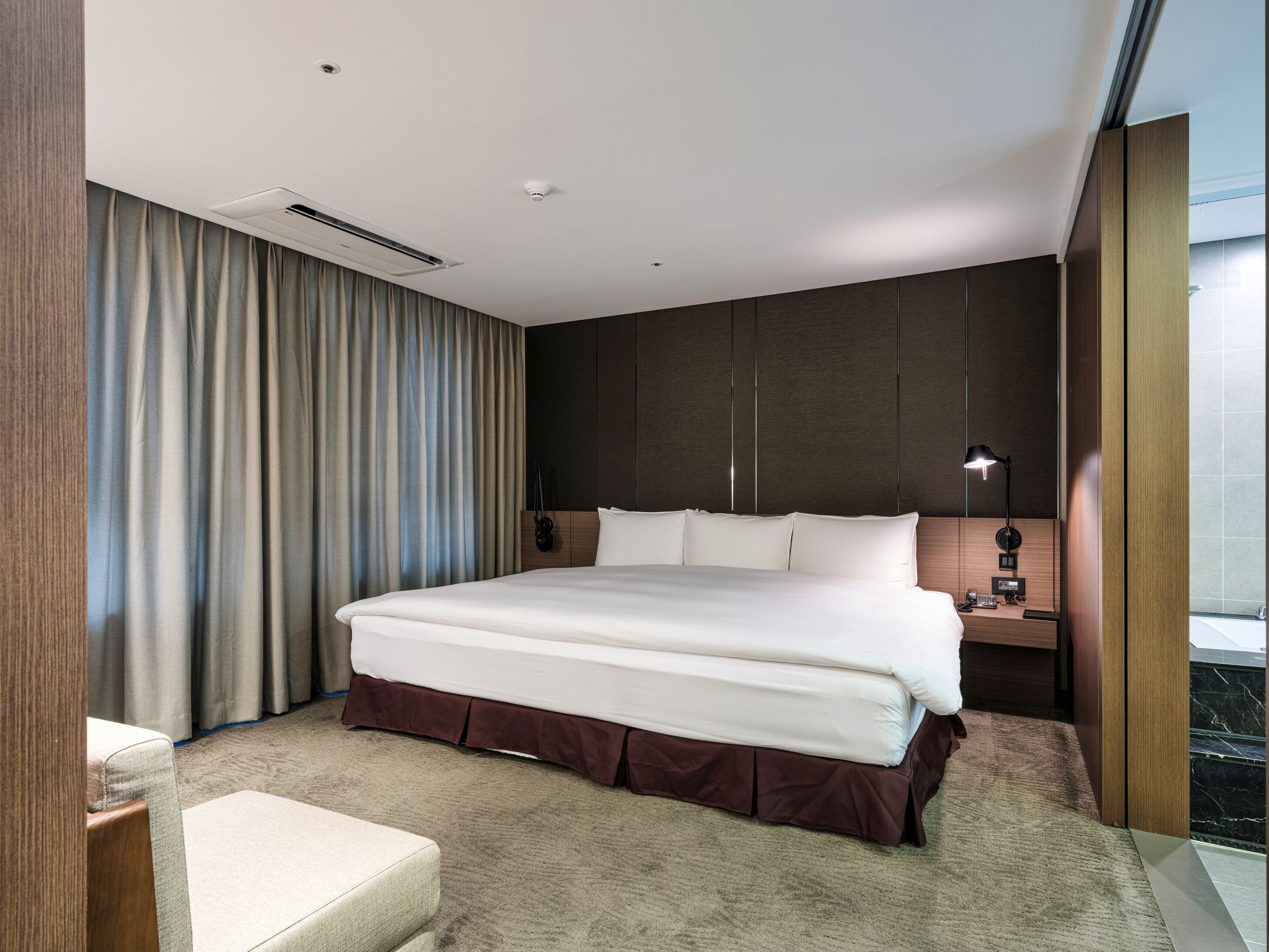 A modern hotel room with a large bed, a bedside table with a lamp, a view of the city through a large window, and a built-in air conditioning unit.