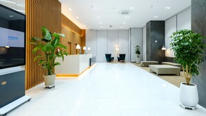 Un área de recepción moderna de hotel con un mostrador de madera, dos sillas, una planta en maceta y una gran pantalla de televisión que muestra el nombre del hotel.