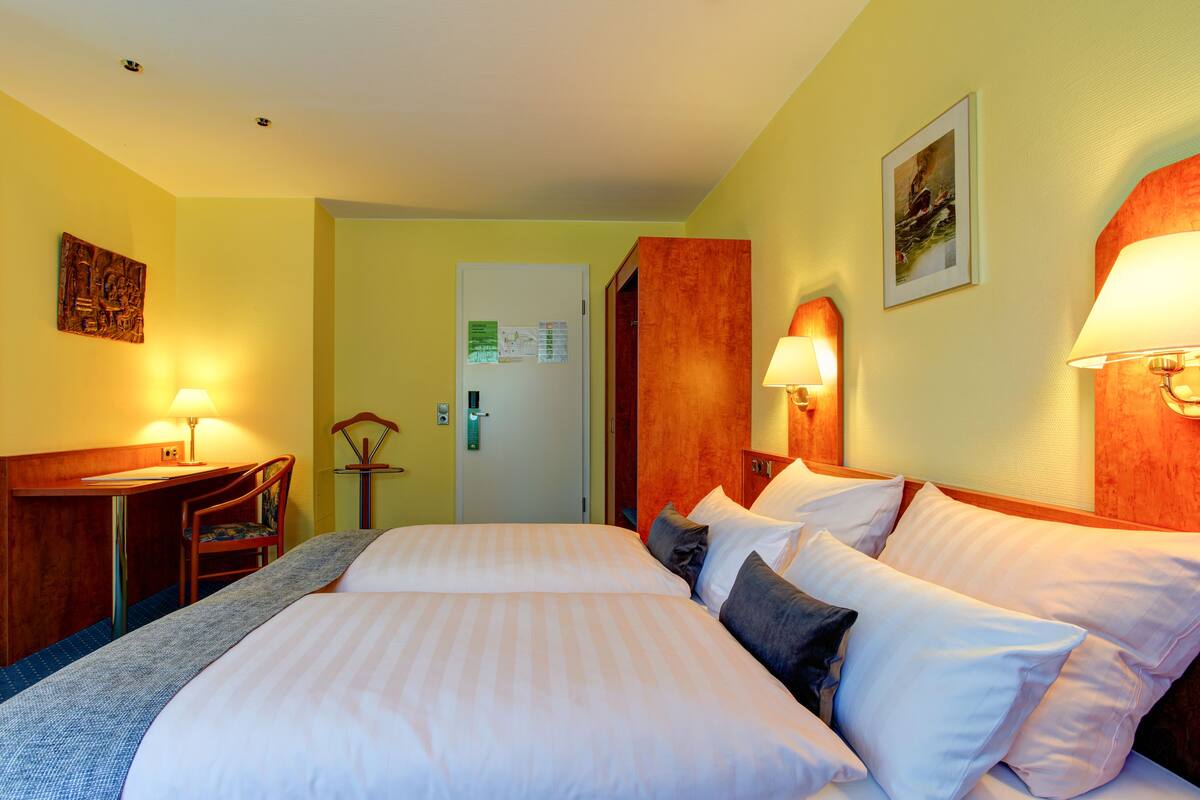 A hotel room with two beds, a desk with a chair, and two wall-mounted lamps.