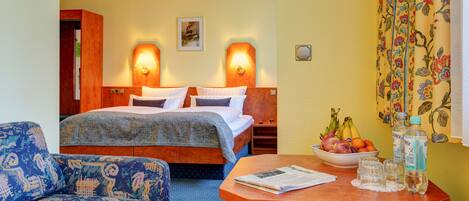 A hotel room with a bed, bedside tables, a sofa, a small table with a newspaper, a bowl of fruit, and a bottle of water.
