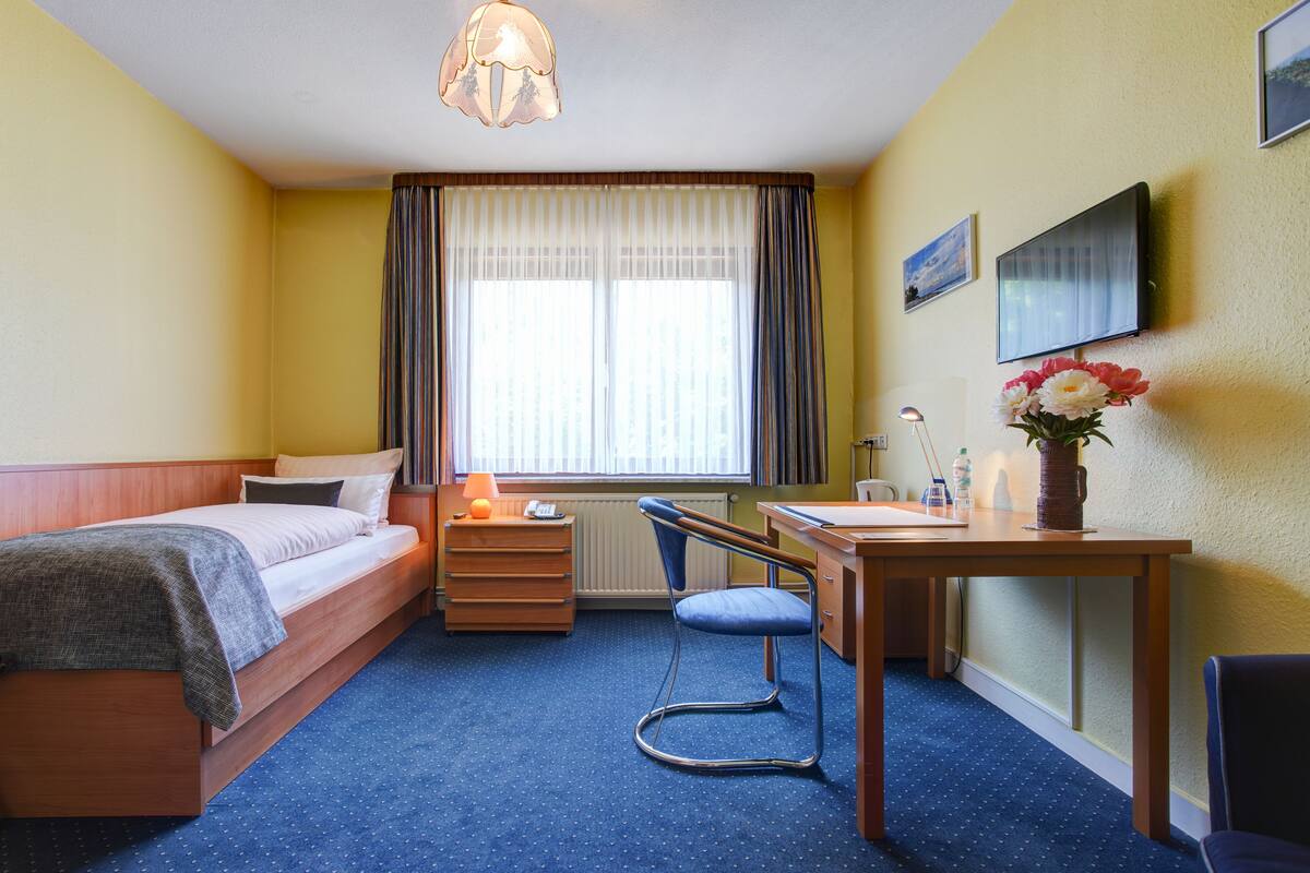 A hotel room with a bed, a desk, a chair, a lamp, a vase with flowers, a television, and a window with curtains.