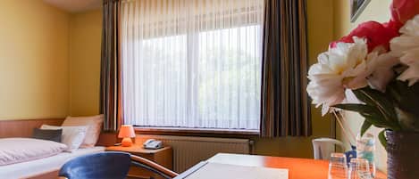 Une chambre d’hôtel avec un lit, un bureau et une chaise, une fenêtre avec des rideaux et un vase rempli de fleurs.
