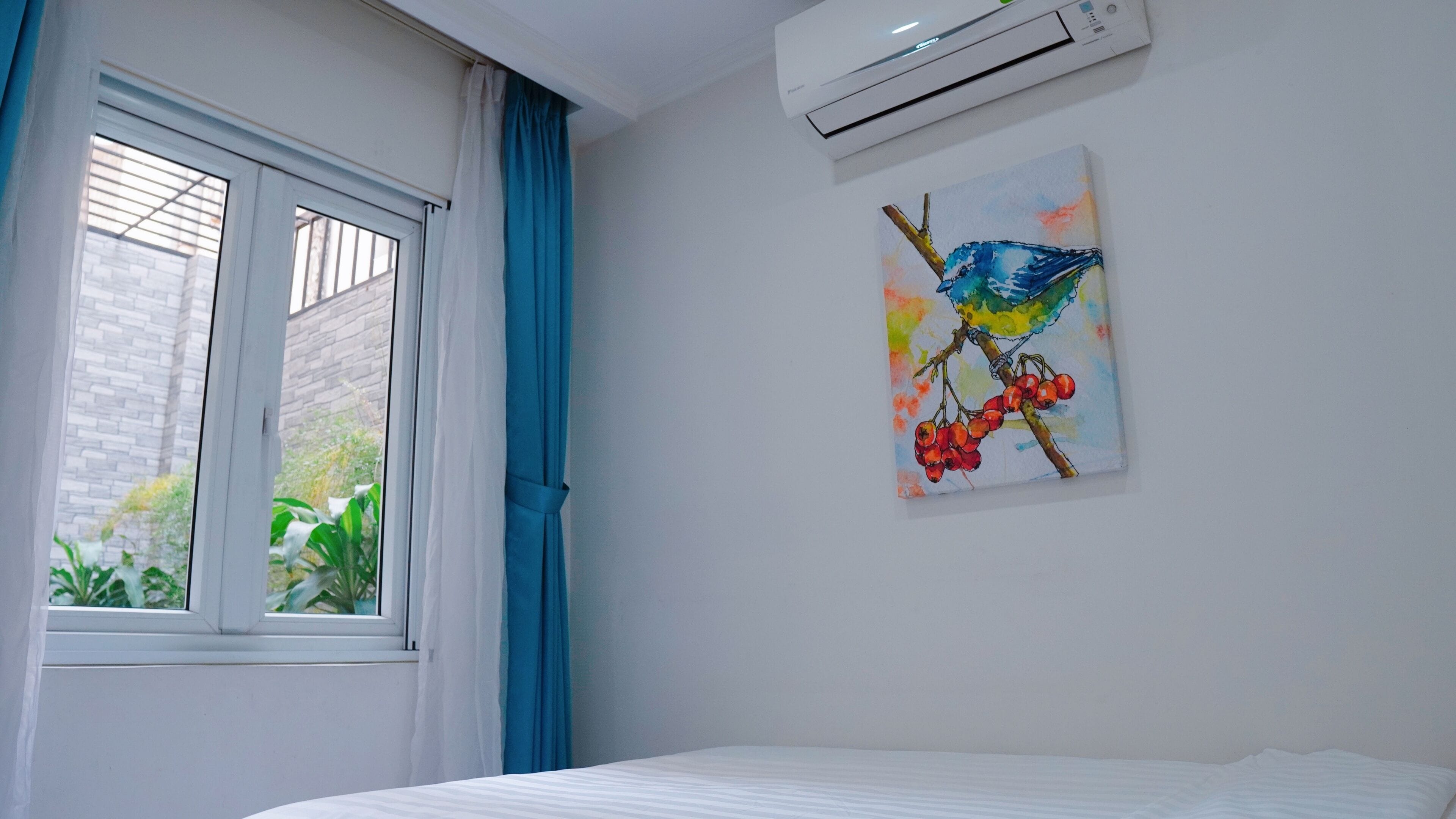 A bedroom with a bed, a window with curtains, an air conditioner, and a wall-mounted painting of a bird.