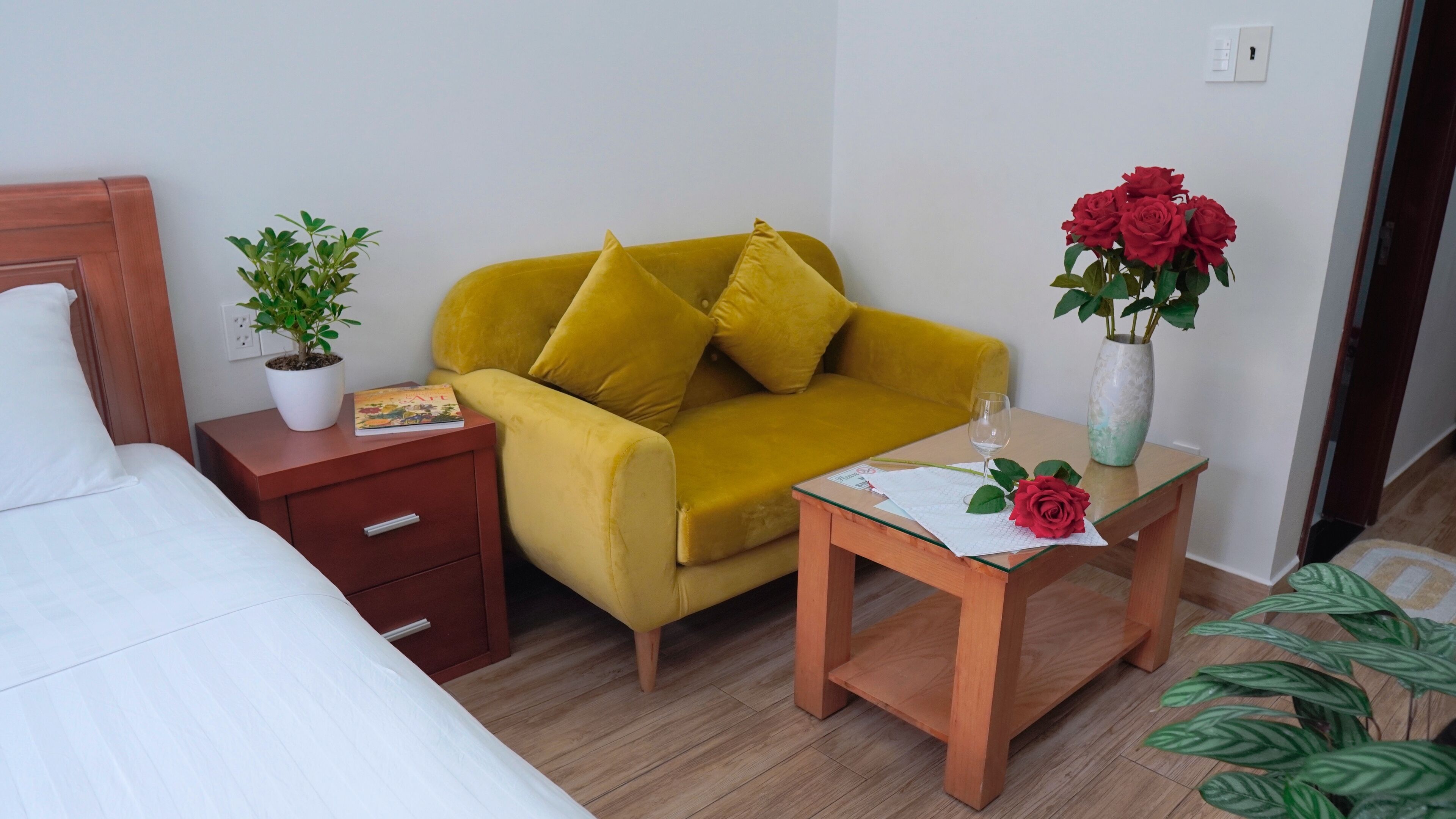 Ein Hotelzimmer mit einem Bett, einem gelben Sofa, einem Nachttisch aus Holz mit einer Pflanze, einem kleinen Tisch mit einer Vase roter Rosen und einem Glas Wein.