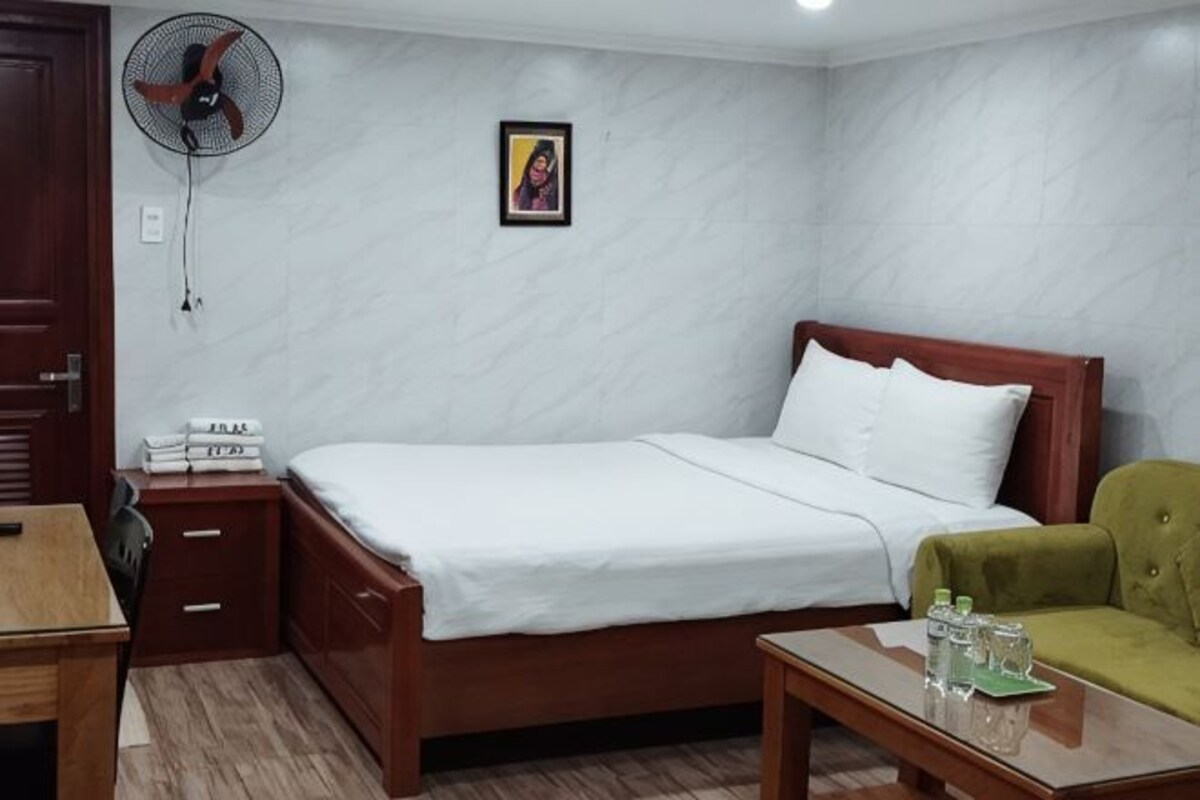 A bedroom with a bed, bedside table, fan, and a green chair.