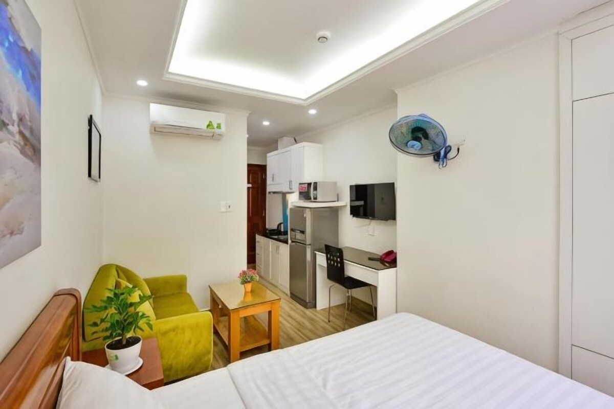 Een hotelkamer met een bed, een bureau, een stoel, een kleine tafel met een plant en een kitchenette op de achtergrond.