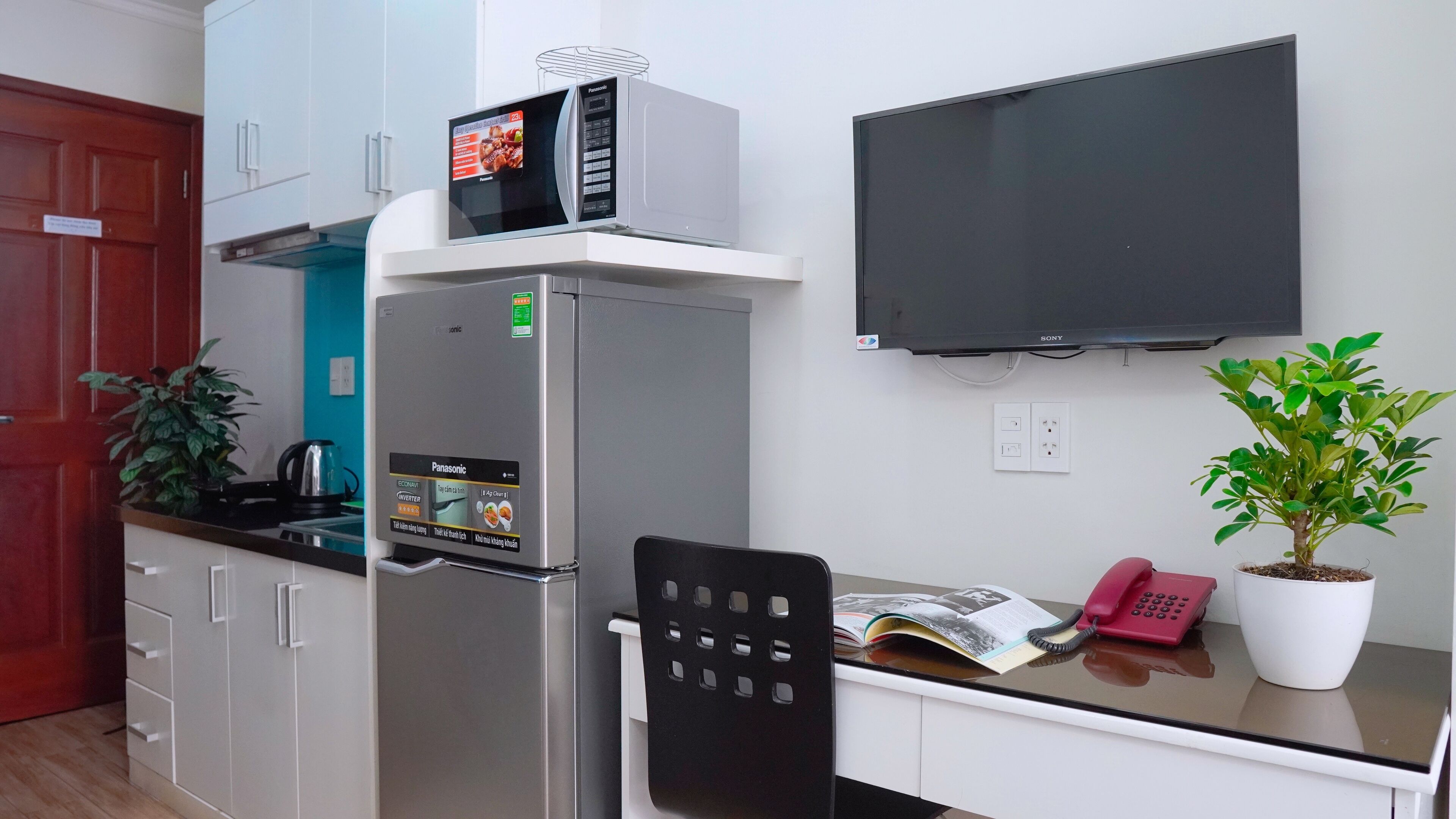 Una kitchenette compacta con microondas integrado, refrigerador y televisor de pantalla plana montado en la pared.