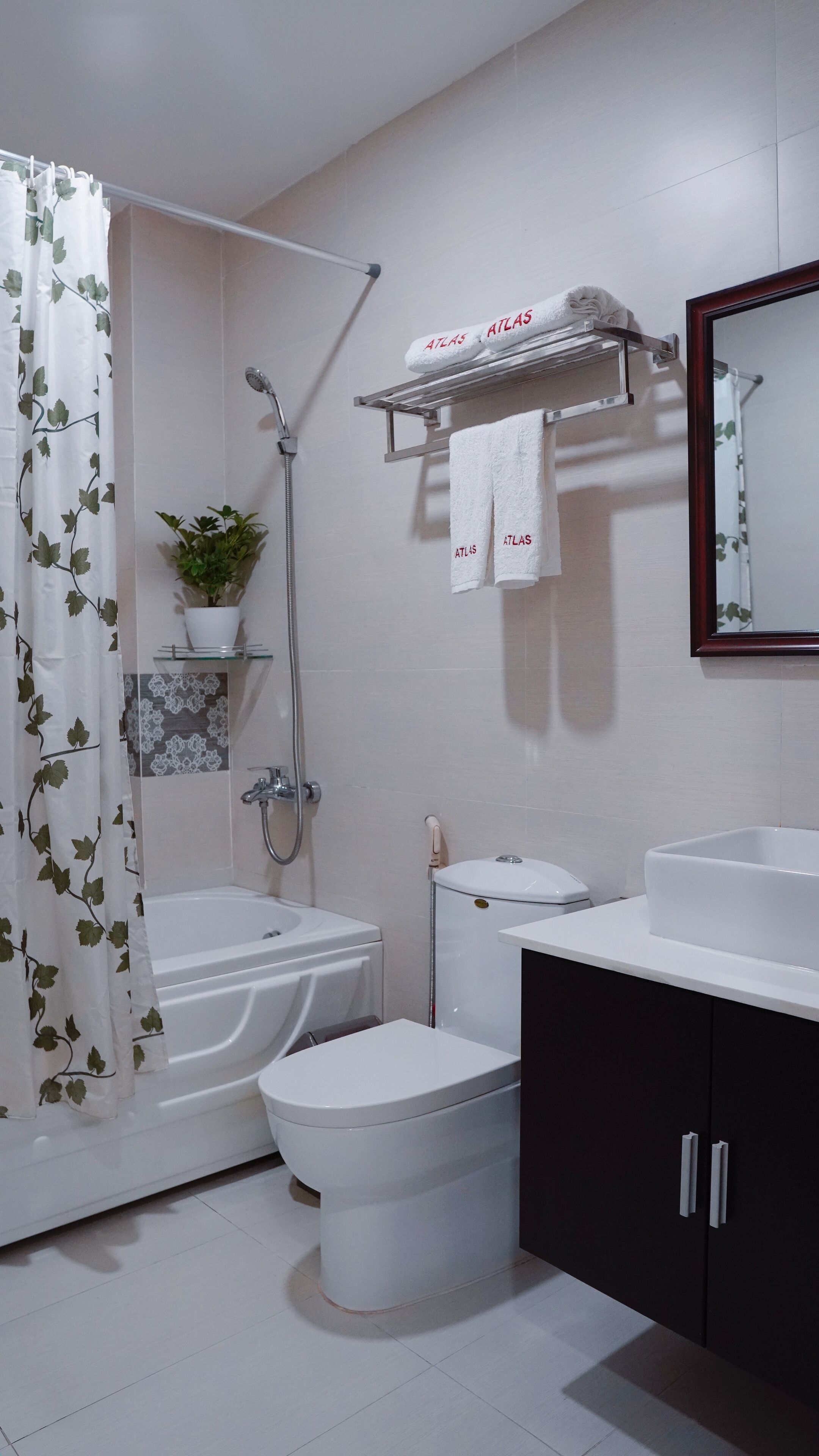 Un baño moderno con ducha, inodoro y lavamanos. Hay un perchero con toallas y una planta en maceta sobre una estante.
