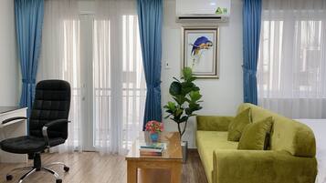 A hotel room with a black office chair, a green sofa, a wooden side table with books and a vase, and a large window with blue curtains.