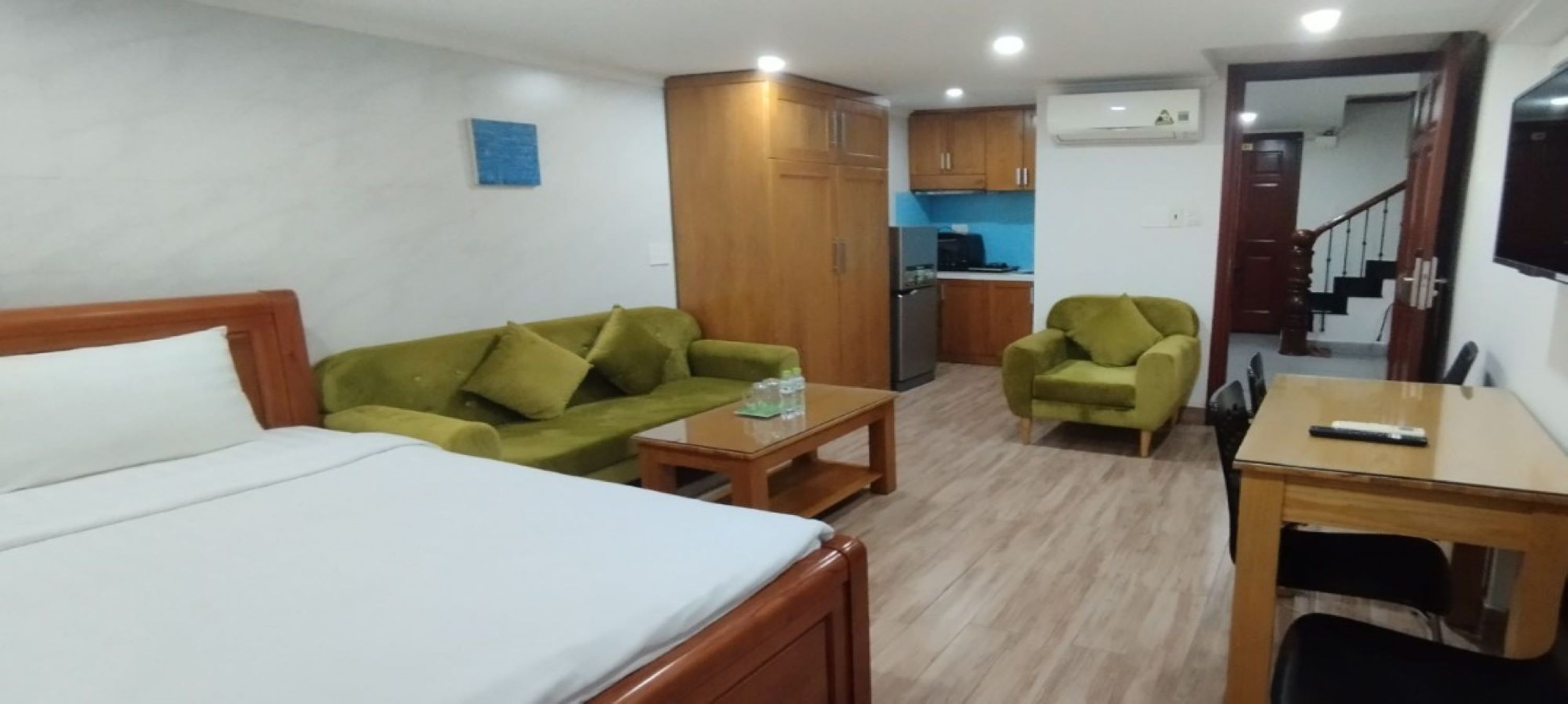 Une chambre d’hôtel avec un lit, un canapé vert, une petite table, un fauteuil vert et, au fond, une kitchenette.
