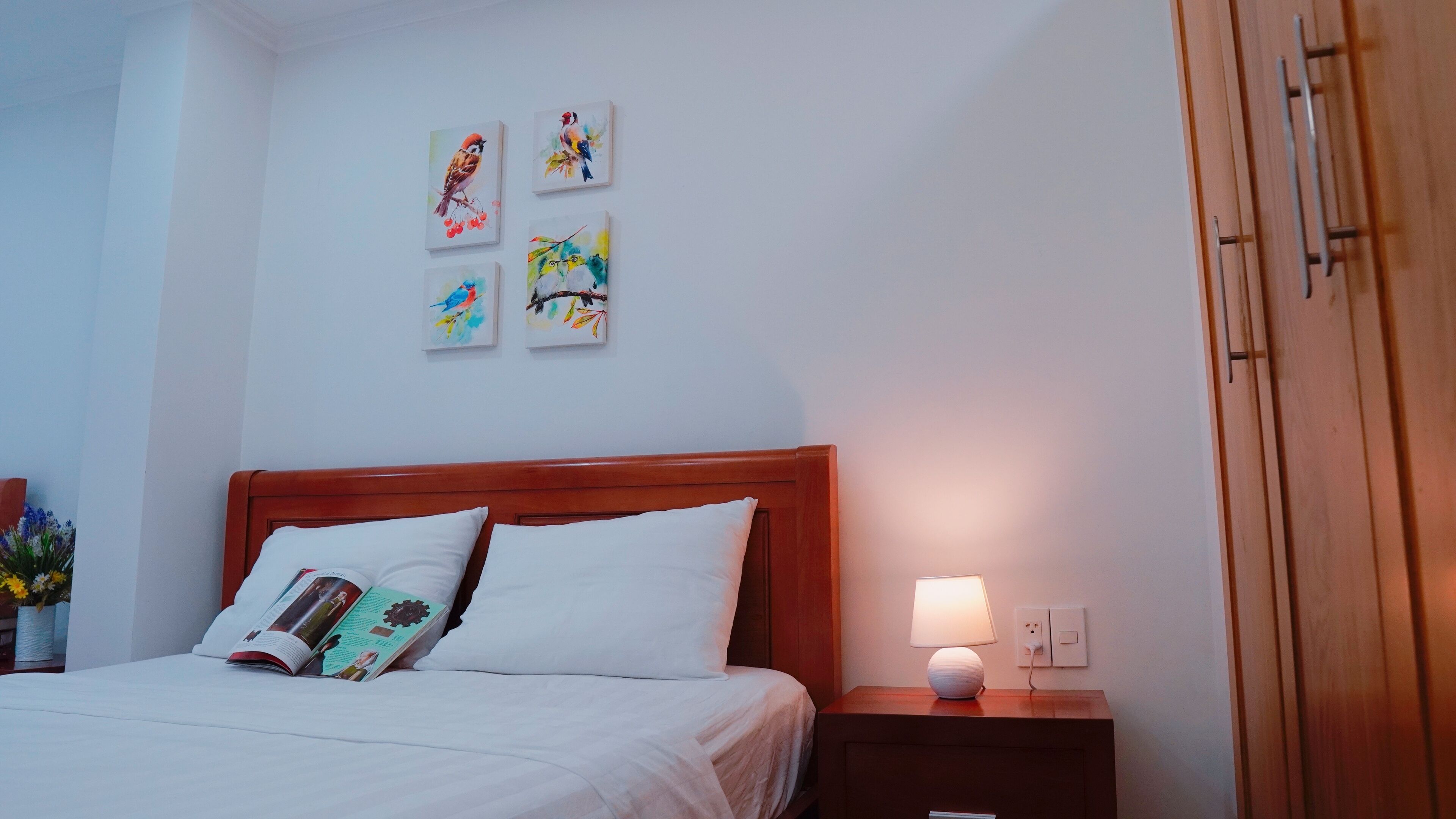 Una cama bien hecha con sábanas y almohadones blancos, una mesita de noche con un libro y una lámpara, y cuatro cuadros de pájaros en la pared.