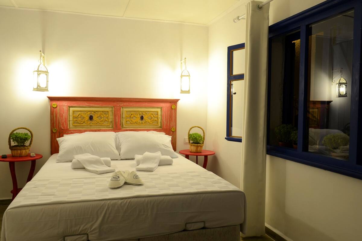A neatly made bed with white linens and towels, two bedside tables with lamps, a window with a view, and a red-framed headboard.