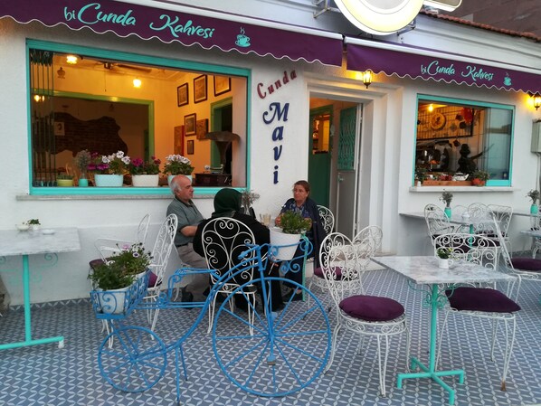 Ein Straßencafé mit Metallstühlen und -tischen, Topfpflanzen und einem Schild mit der Aufschrift 'bi Cunda Kahvesi'.