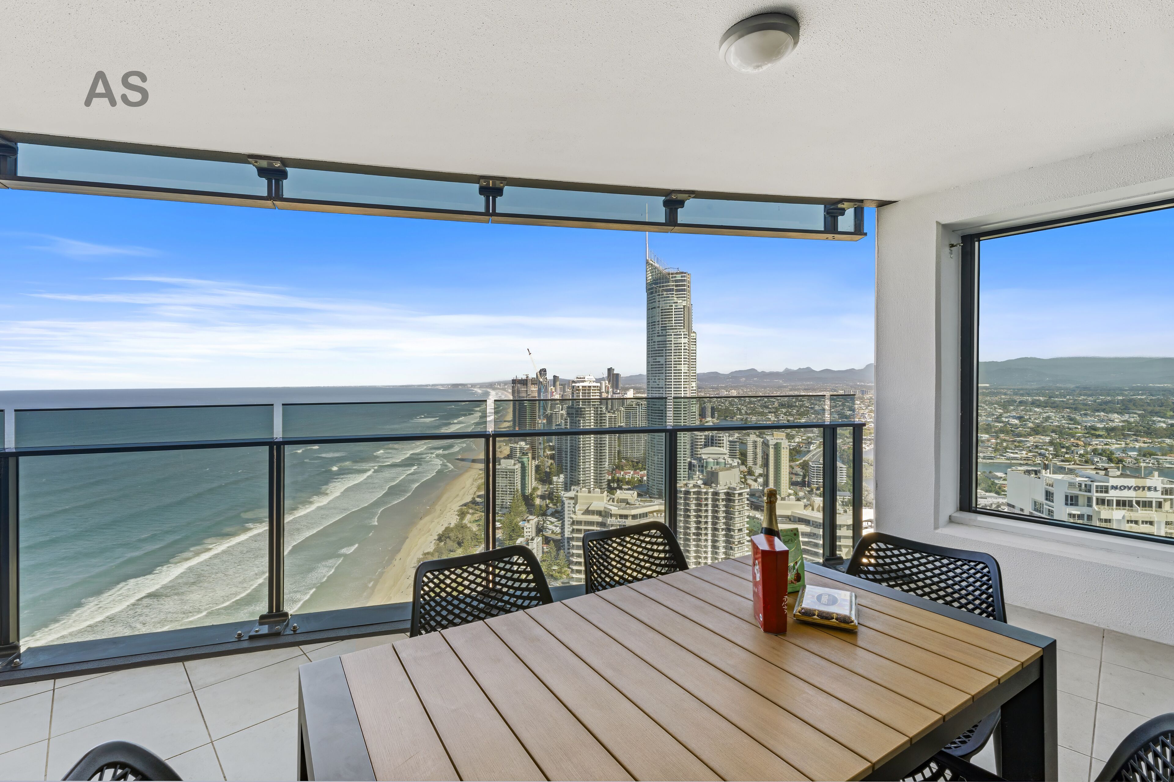 A balcony with a wooden table and chairs, overlooking a beach and cityscape with a tall building in the distance.