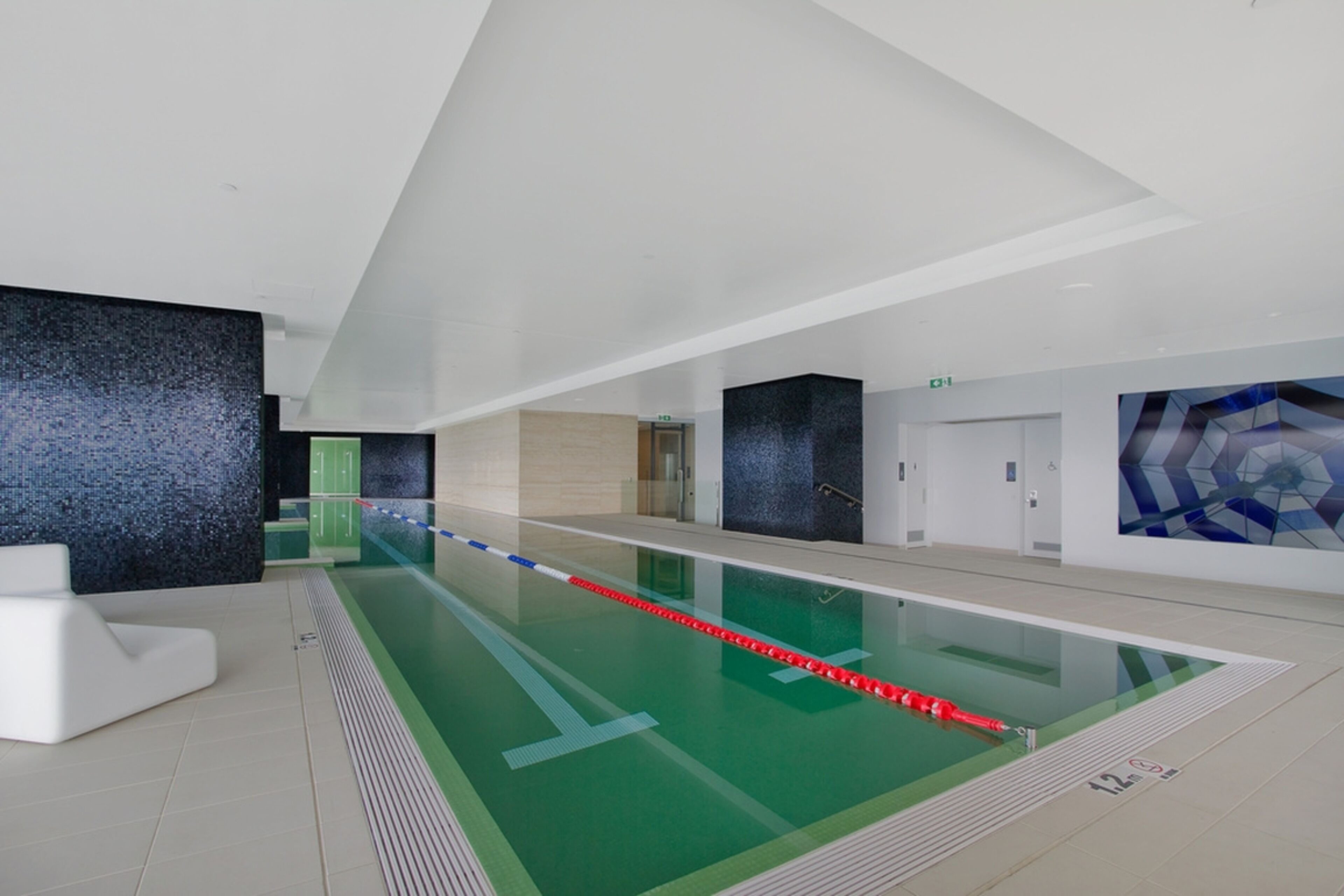 Une piscine intérieure moderne dotée d’une ligne de nage bien délimitée, d’un espace d’accueil blanc et d’un motif géométrique sur le mur.